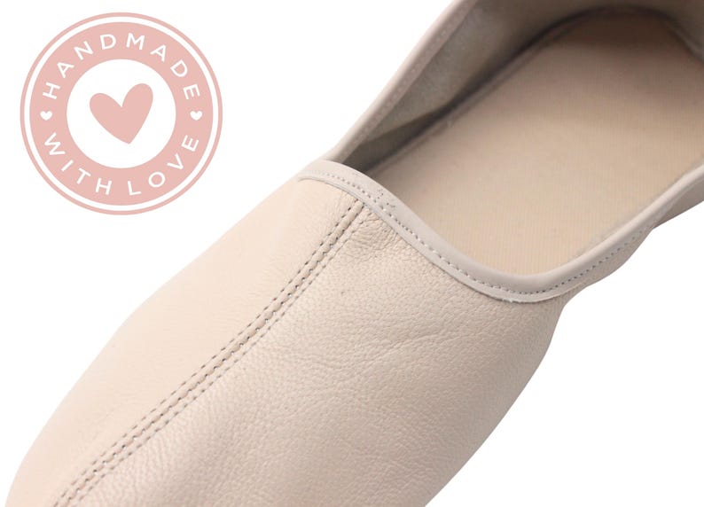 Pu&ograve; includere: Primo piano di una ballerina color crema. La scarpa &egrave; realizzata in pelle e presenta una cucitura cucita al centro. Una grafica circolare rosa con le parole "HANDMADE WITH LOVE" circonda un cuore rosa.