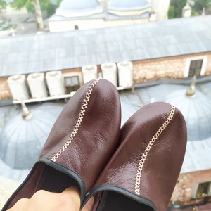 May include: A pair of brown leather slippers with a decorative stitch detail. The slippers have a black trim around the edges.