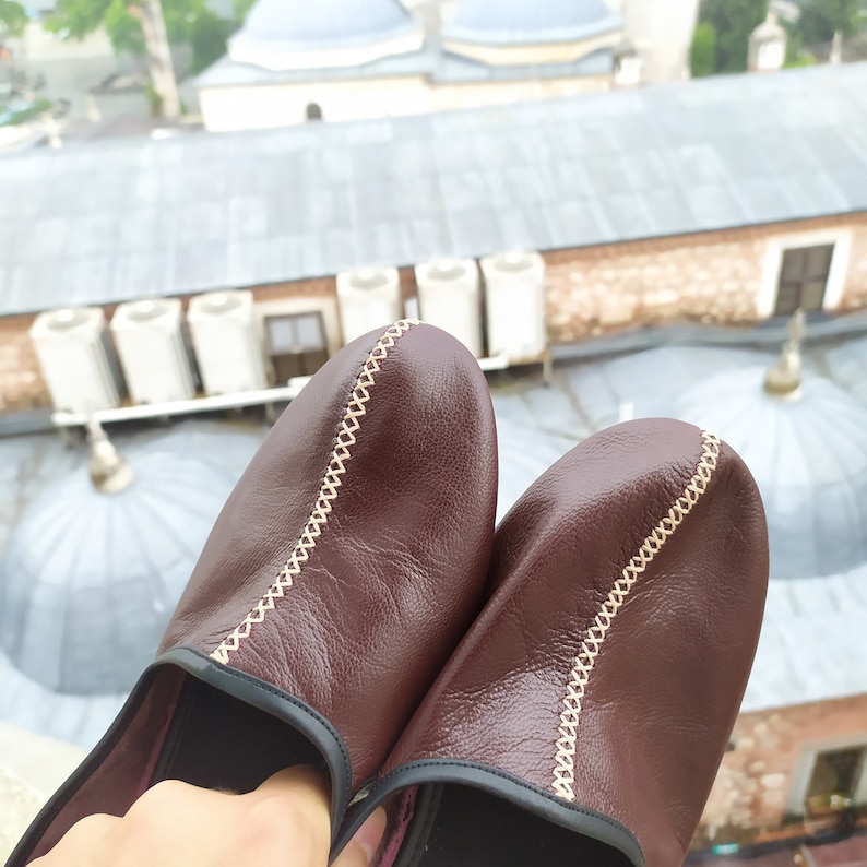 May include: A pair of brown leather slippers with a decorative stitch detail. The slippers have a black trim around the edges.