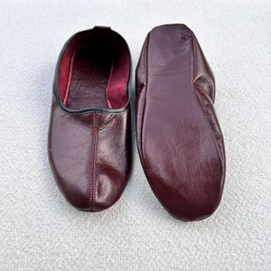 Puede incluir: Un par de zapatillas de cuero burdeos con un ribete negro alrededor de la abertura. Las zapatillas est&aacute;n hechas de cuero suave y flexible y tienen un ajuste c&oacute;modo.