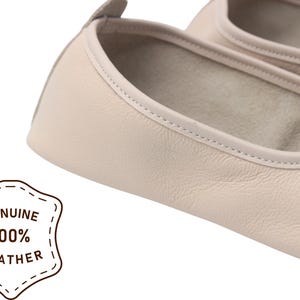 Pu&ograve; includere: Primo piano di un paio di ballerine in pelle beige chiaro. Le scarpe hanno la punta arrotondata e un bordo cucito. L'immagine include un distintivo che recita "GENUINE 100% LEATHER".