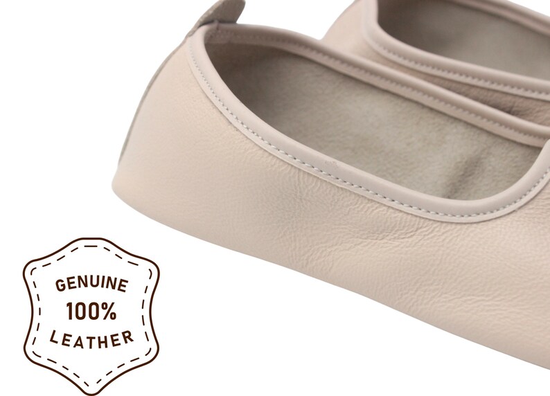 Pu&ograve; includere: Primo piano di un paio di ballerine in pelle beige chiaro. Le scarpe hanno la punta arrotondata e un bordo cucito. L'immagine include un distintivo che recita "GENUINE 100% LEATHER".