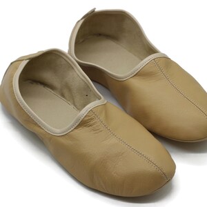 May include: A pair of tan leather slippers with a white trim around the opening. The slippers have a pointed toe and a simple design.