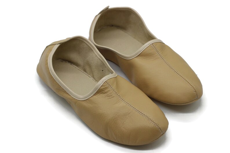 May include: A pair of tan leather slippers with a white trim around the opening. The slippers have a pointed toe and a simple design.
