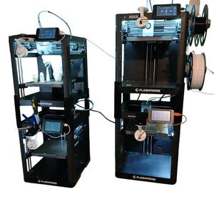 May include: Two black Flashforge 3D printers with multiple shelves, screens, and attached filament spools. One printer has the text "AD5X" on the top. The printers are connected by a white cable. The printers are on a white background.