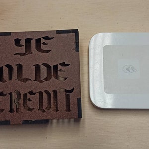May include: A wooden sign with the words "Ye Olde Credit" carved into it. The sign is brown with black trim and is sitting on a wooden surface next to a white square-shaped credit card reader.