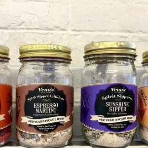 May include: Four glass jars with gold lids filled with different ingredients. Each jar has a label with the text "Vena's Spirit Sippers" and a description of the flavor. The flavors are Sweet Heat, Espresso Martini, Sunshine Sipper, and Blushing Grapefruit.