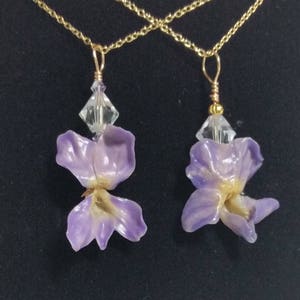 Puede incluir: Dos collares dorados, cada uno con un colgante de una flor de color lila claro y una cuenta de cristal transparente. Los colgantes de flores tienen un aspecto delicado y natural, con un acento dorado en el centro.