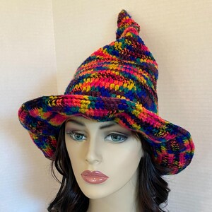 May include: A colorful, handmade crochet hat with a wide brim and a pointed top. The hat features a vibrant mix of rainbow colors, including pink, blue, yellow, and purple. The hat is designed with a textured stitch.