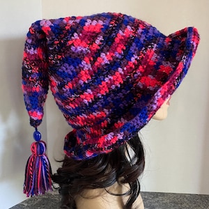 May include: A colorful, handmade crochet hat in shades of red, pink, purple, and blue. The hat features a long, pointed tip with a blue bead and a tassel. The brim is wide and the hat is designed to be worn.