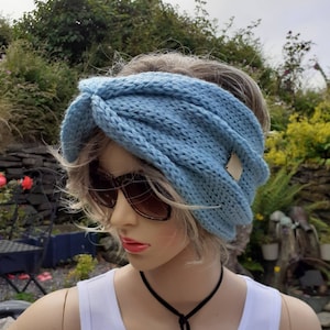 May include: A light blue knitted headband with a twist detail. The headband has a small brown leather tag attached to it.