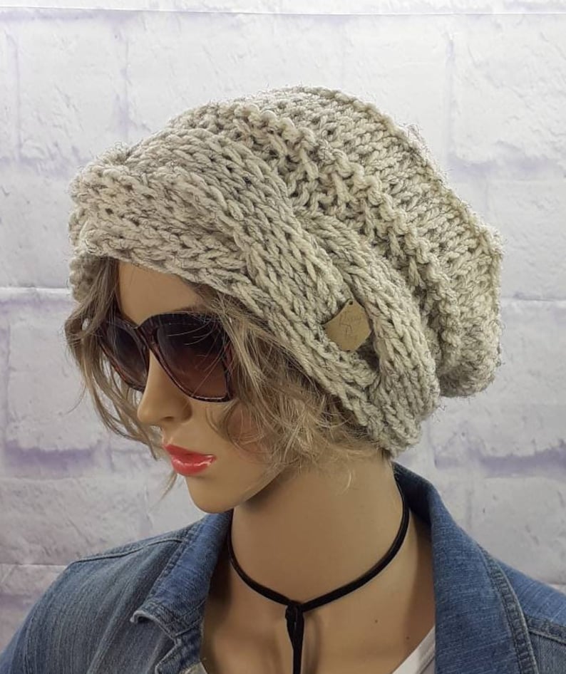 May include: A slouchy, light gray knitted beanie with a brown button detail. The beanie is made of a chunky yarn and has a relaxed fit.