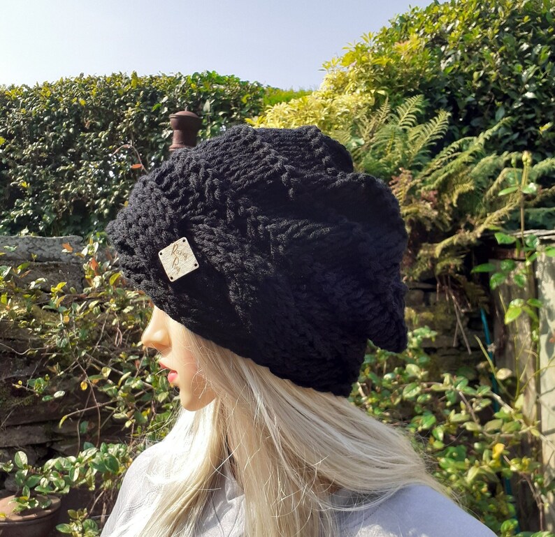 May include: A black knitted beanie with a small wooden tag that says "Rainy Day". The beanie is being worn by a person with blonde hair.
