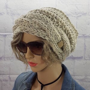 May include: A gray knitted beanie with a unique twisted design. The beanie has a small brown leather tag with a letter on it.