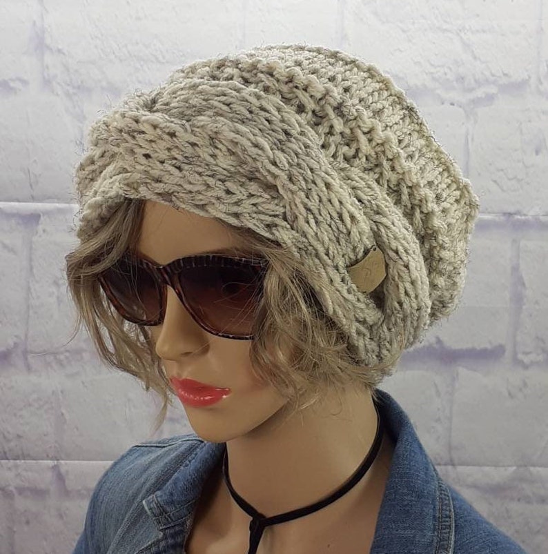 May include: A gray knitted beanie with a unique twisted design. The beanie has a small brown leather tag with a letter on it.