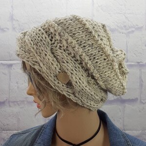 May include: A slouchy, beige knitted beanie with a cable knit pattern. The beanie has a small, brown leather tag attached to the side.