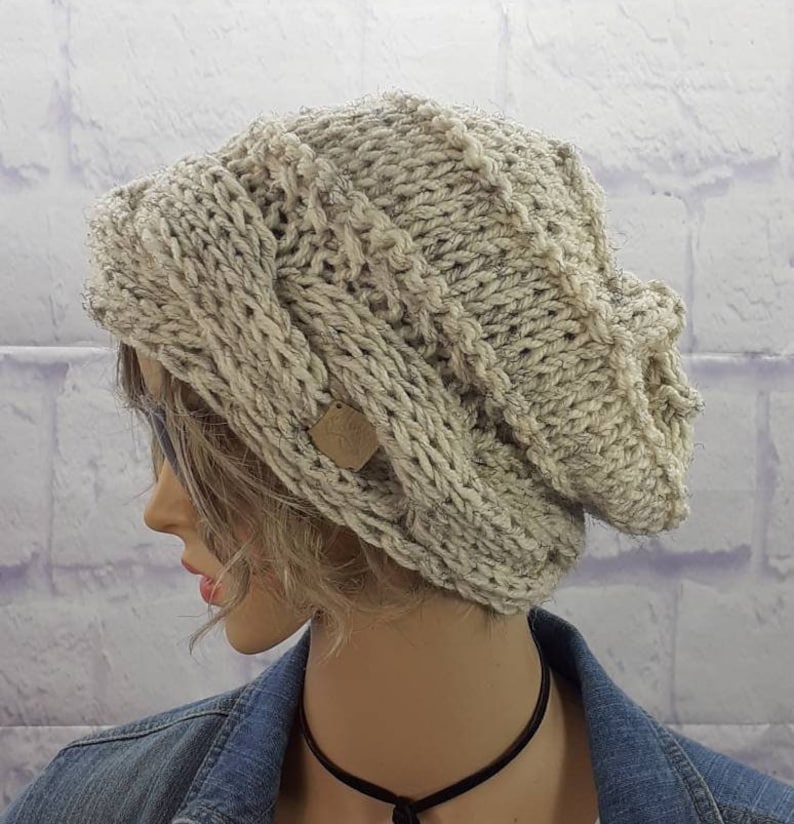 May include: A slouchy, beige knitted beanie with a cable knit pattern. The beanie has a small, brown leather tag attached to the side.
