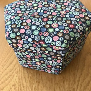 Peut inclure: Une boîte de rangement hexagonale avec un fond bleu et un motif de boutons colorés. La boîte est en tissu et a une texture douce et matelassée.