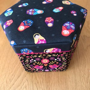 Pode incluir: Uma caixa hexagonal de tecido com fundo preto e um padrão de bonecas russas coloridas. A caixa tem um padrão floral preto e branco na parte inferior.