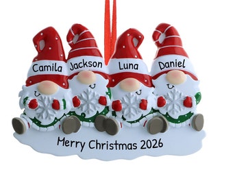 Op de afbeelding: Een kerstversiering met vier kerstmannetjes met rode mutsen en witte baarden. Elk kerstmannetje houdt een sneeuwvlok vast en heeft een naam op zijn muts: Camila, Jackson, Luna en Daniel. De versiering bevat ook de tekst "Prettige Kerst 2026".