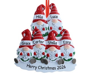 Op de afbeelding: Een kerstversiering met zes kerstmannetjes met rode mutsen en witte baarden. Elk kerstmannetje heeft een naam op zijn muts: Mila, Luca, Grace, Carter, Stella en Ezra. De versiering bevat ook de tekst "Prettige Kerst 2026".