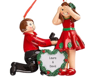 May include: A Christmas ornament showing a marriage proposal scene. A man in a red jacket kneels, offering a ring. A woman in a red dress covers her eyes. The ornament includes the date 08/26/2026 and the names Laura & David.