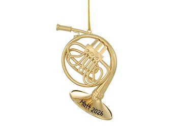 Puede incluir: Adorno navideño dorado con forma de trompa francesa, con una cuerda dorada para colgar. El adorno tiene el texto "Matt 2026" grabado en la campana. El instrumento es un instrumento musical.