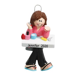 Puede incluir: Un adorno navideño personalizado que representa a una mujer con cabello castaño sentada en una mesa con esmalte de uñas, una lima de uñas y cortaúñas. La mesa es blanca y tiene el texto "Jennifer 2020" escrito en ella.