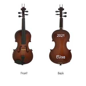 Puede incluir: Un violín marrón en miniatura, vistas frontal y trasera. La parte delantera muestra las cuerdas y el puente. La parte trasera tiene el texto "2021" y "Elisa". Es un objeto de colección musical.