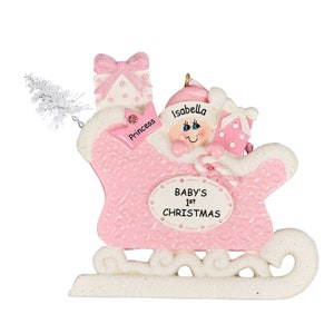 May include: Pink personalized Christmas ornament featuring a baby girl in a sleigh with presents. The ornament reads "Baby's 1st Christmas" and is decorated with glitter and a snowflake design.