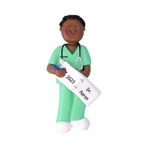 May include: A green scrubs ornament with a stethoscope, pen, and notepad. The notepad says "2023" and "Dr. Aaron".