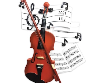 May include: A decorative ornament featuring a reddish-brown violin with a bow, set against a backdrop of musical notes and sheet music. The ornament includes the text "2021 Lilly" on a banner.