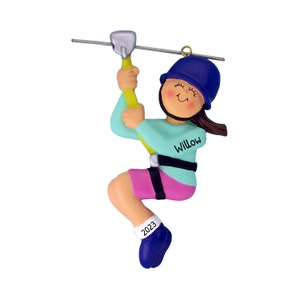 Peut inclure: Une d&eacute;coration de No&euml;l personnalis&eacute;e repr&eacute;sentant une fille en tyrolienne. Elle porte un casque bleu, un t-shirt turquoise avec le nom "Willow" &eacute;crit dessus, un short rose et des chaussures bleues. La d&eacute;coration est dat&eacute;e de "2023".