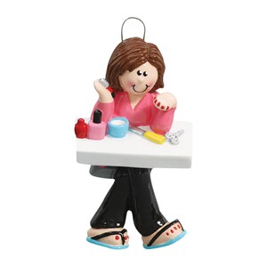 Puede incluir: Un adorno de cer&aacute;mica pintado a mano que representa a una persona sentada en una mesa con esmalte de u&ntilde;as, una lima de u&ntilde;as y corta&uacute;&ntilde;as. La persona lleva una camisa rosa y pantalones negros.