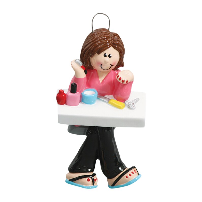 Puede incluir: Un adorno de cer&aacute;mica pintado a mano que representa a una persona sentada en una mesa con esmalte de u&ntilde;as, una lima de u&ntilde;as y corta&uacute;&ntilde;as. La persona lleva una camisa rosa y pantalones negros.
