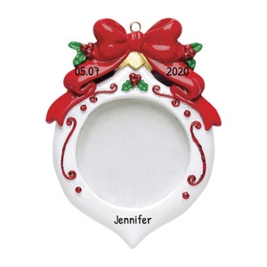 Puede incluir: Adorno navideño blanco con lazo rojo, detalles de acebo y el año 2020. El adorno tiene una abertura circular para una foto y el nombre "Jennifer" está impreso en la parte inferior.