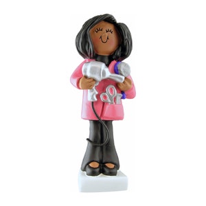 May include: A figurine of a hairstylist, featuring a smiling face and black hair. The figure wears a pink coat over a black top and pants, holding a silver hairdryer and a blue hairbrush. A pair of scissors is in the coat pocket. The figurine stands on a white base.