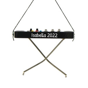 May include: A black ornament shaped like a keyboard with the text "Isabella 2022" in white. The ornament has a series of colored buttons and gray knobs. It is suspended by a black string and has silver legs.