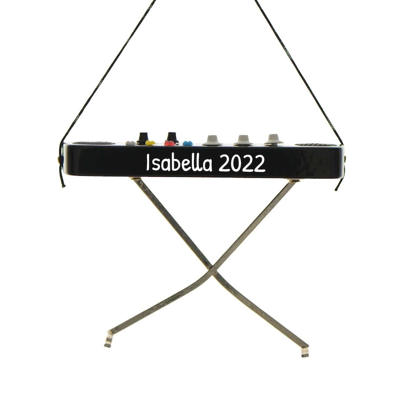 May include: A black ornament shaped like a keyboard with the text "Isabella 2022" in white. The ornament has a series of colored buttons and gray knobs. It is suspended by a black string and has silver legs.