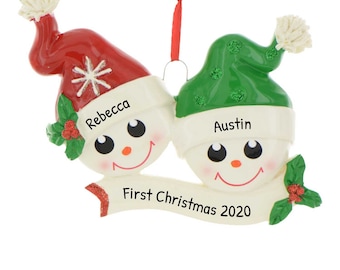 May include: A personalised Christmas bauble featuring two snowmen with red and green hats. The snowmen are holding a banner that reads "First Christmas 2020". The snowmen are named "Rebecca" and "Austin".