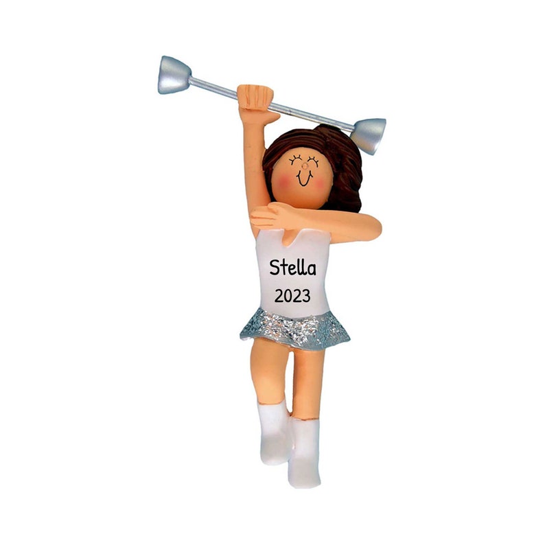 May include: A figurine of a baton twirler in a white top and silver skirt, holding a silver baton. The figurine has brown hair and is wearing white boots. The name "Stella" and the year "2023" are printed on the top.