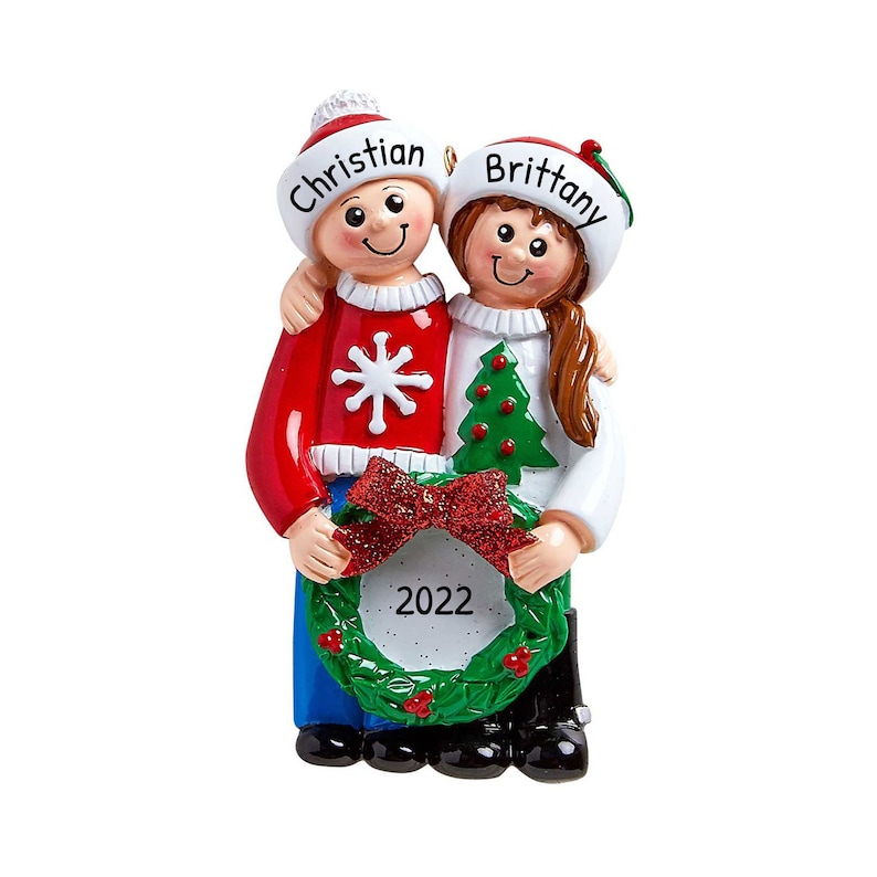 Puede incluir: Adorno navide&ntilde;o personalizado con una pareja que lleva su&eacute;teres rojos y blancos, sosteniendo una corona verde con un lazo rojo y el a&ntilde;o 2022. El adorno est&aacute; decorado con copos de nieve y un peque&ntilde;o &aacute;rbol de Navidad. Los nombres "Christian" y "Brittany" est&aacute;n escritos en sus gorros.