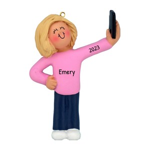 May include: A personalized Christmas ornament featuring a blonde woman wearing a pink shirt and blue pants, taking a selfie with a black phone. The ornament is inscribed with the name "Emery" and the year "2023".