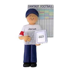 May include: A white and blue personalised ornament of a person wearing a blue baseball cap and holding a laptop with the year 2023 on the screen. The person is wearing a white shirt with the name Jaxson on it. The ornament is next to a fantasy football score sheet.