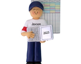 May include: A white and blue personalized ornament of a person wearing a blue baseball cap and holding a laptop with the year 2023 on the screen. The person is wearing a white shirt with the name Jaxson on it. The ornament is next to a fantasy football score sheet.