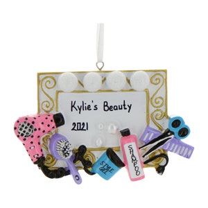 May include: A white framed ornament with gold trim and a mirror with light bulbs. The ornament has the text "Kylie's Beauty 2021" and features a pink hair dryer, a purple hair brush, a blue hair gel container, a pink shampoo bottle, a purple hair straightener, and a pair of blue scissors.