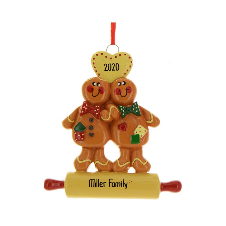 Peut inclure: Une d&eacute;coration de No&euml;l en forme de couple de pain d'&eacute;pices avec un c&oelig;ur jaune sur le dessus qui dit "2020". Les personnages en pain d'&eacute;pices se tiennent la main et se tiennent sur un rouleau &agrave; p&acirc;tisserie jaune qui dit "Miller Family".