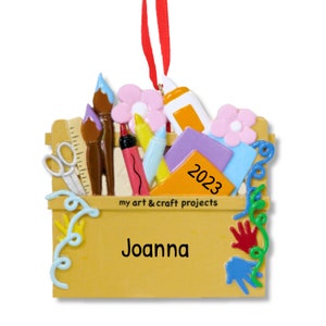 May include: A yellow wooden craft box filled with art supplies, including paintbrushes, scissors, markers, and paper. The box has the text "my art & craft projects" and "Joanna" on the front. The year 2023 is written on a piece of orange paper inside the box.