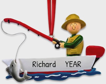 May include: A hanging ornament featuring a figure in a yellow vest and tan hat sitting in a white boat with a red fishing rod. The boat has the text Richard and YEAR printed on the side. A silver fish hangs from the line, and a red ribbon is attached.