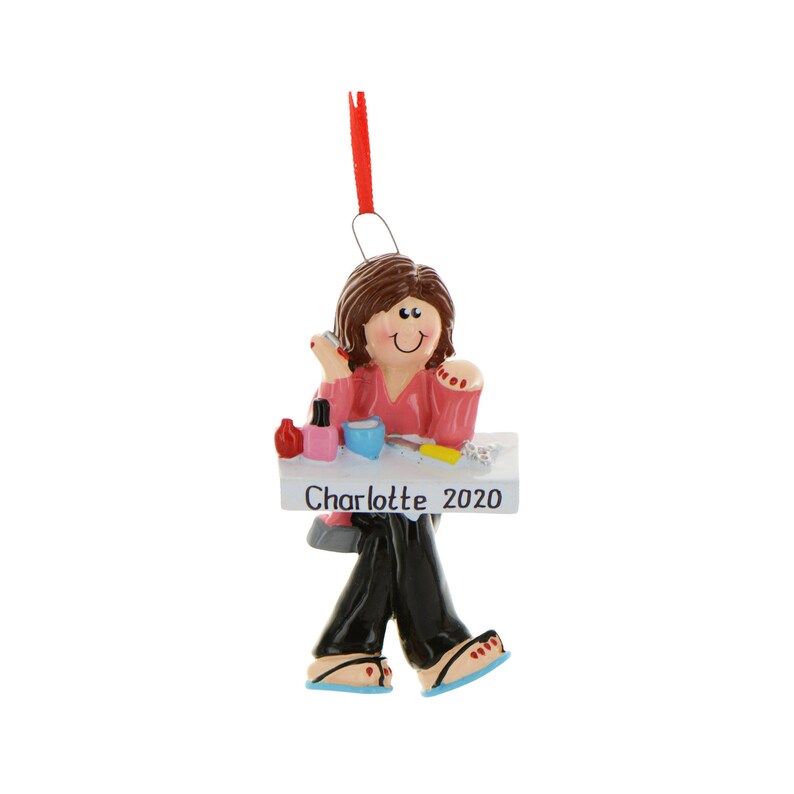 Puede incluir: Un adorno navide&ntilde;o personalizado que representa a una mujer pint&aacute;ndose las u&ntilde;as en una mesa. El adorno es blanco con el nombre "Charlotte" y el a&ntilde;o "2020" escrito en &eacute;l.